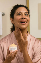 A woman applying The Rejuvenating Neck Firming Cream in a bathroom, highlighting the cream's moisturizing benefits.